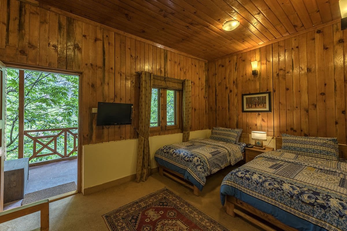 Quarto de cabana de madeira com duas camas, uma TV e uma janela grande com vista para árvores.