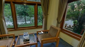 A room with a wooden chair, a glass table with water bottles, and large windows offering a view of greenery. - Arcadian Riverside (Balakot)