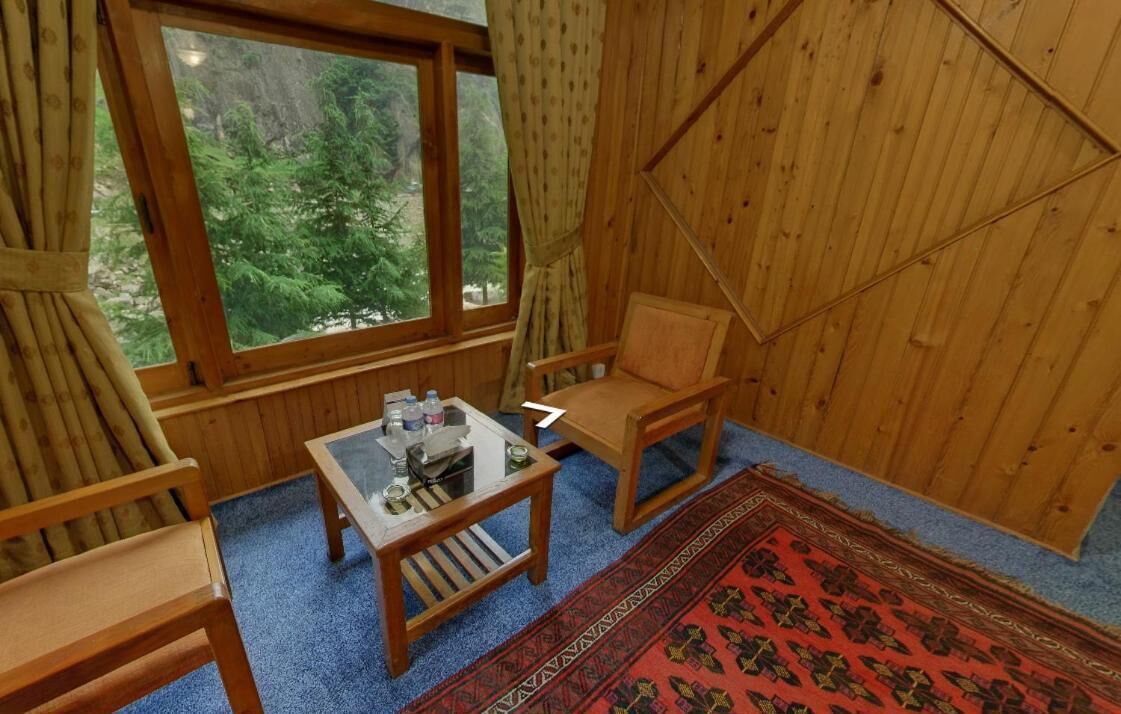 Een gezellige kamer met een groot raam, houten wanden, een klein tafeltje, twee stoelen en een kamerkleed met een patroon.