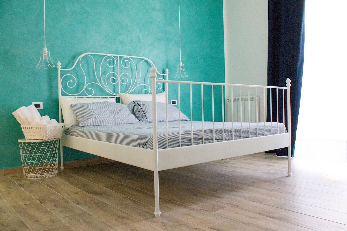 A white metal bed with a decorative headboard, a striped mattress, and a white metal side table with a basket.