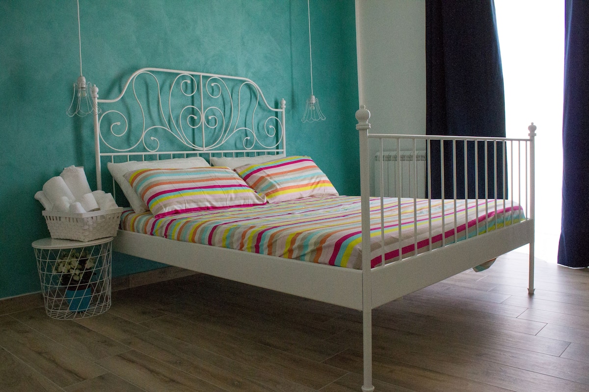 A white metal bed with a colorful striped bedspread and pillows, a white metal headboard, and a white metal nightstand with a basket.