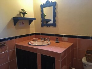 A bathroom with a sink, mirror, and toilet. There is a towel rack and a small shelf with a plant. - Casita El Huipil (Antigua Guatemala)