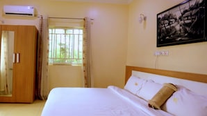 A bedroom with a bed, a window with curtains, an air conditioning unit, and a painting on the wall.