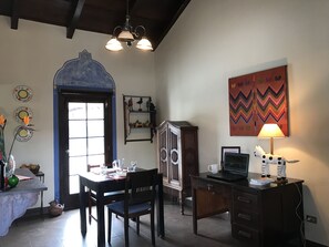 A dining area with a table set for two, a cabinet, a desk with a lamp, and a colorful wall hanging. - Casita Buena Vista (Antigua Guatemala)