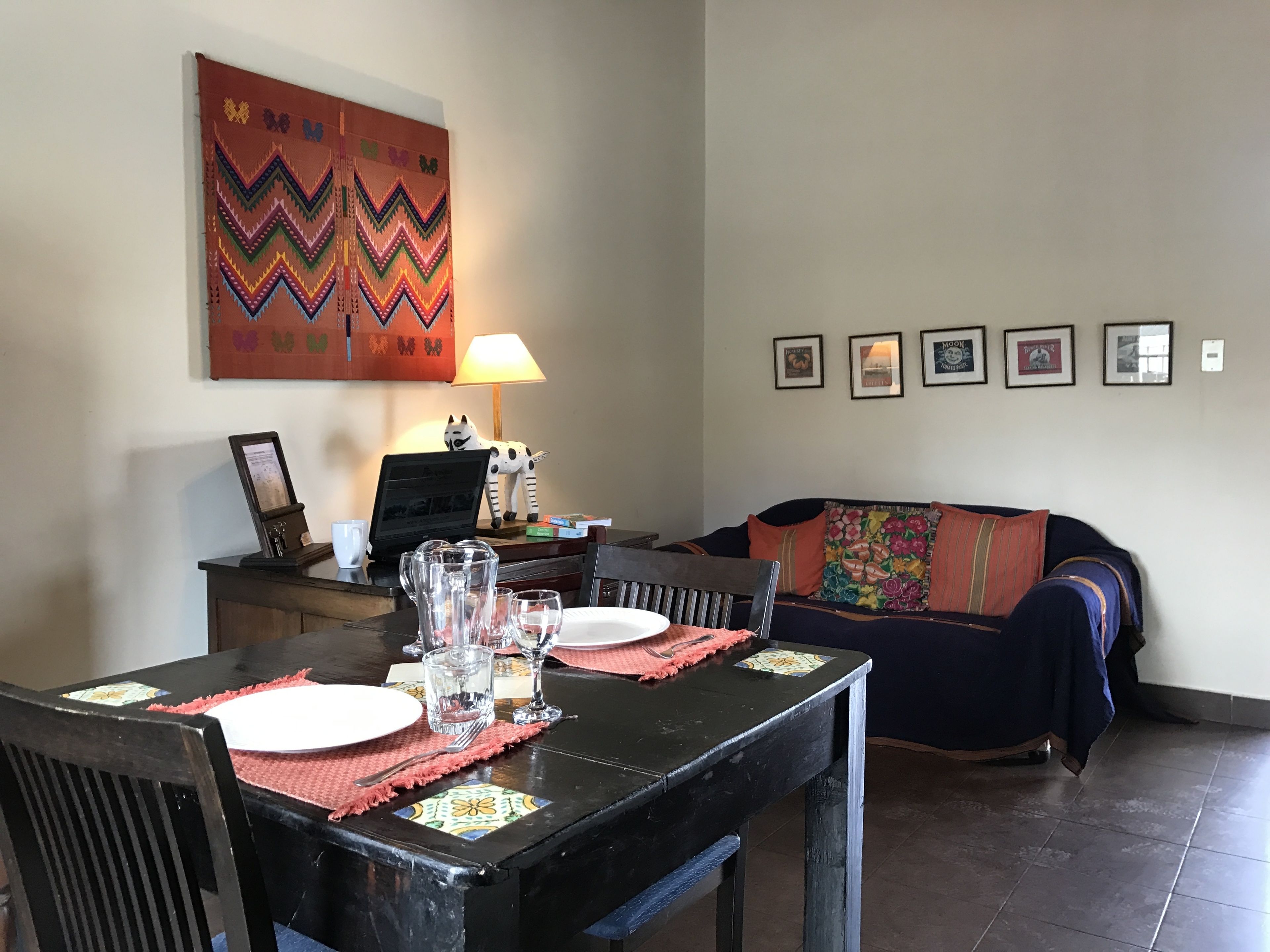 A dining area with a table set for two, a laptop, a lamp, and a sofa in the background.