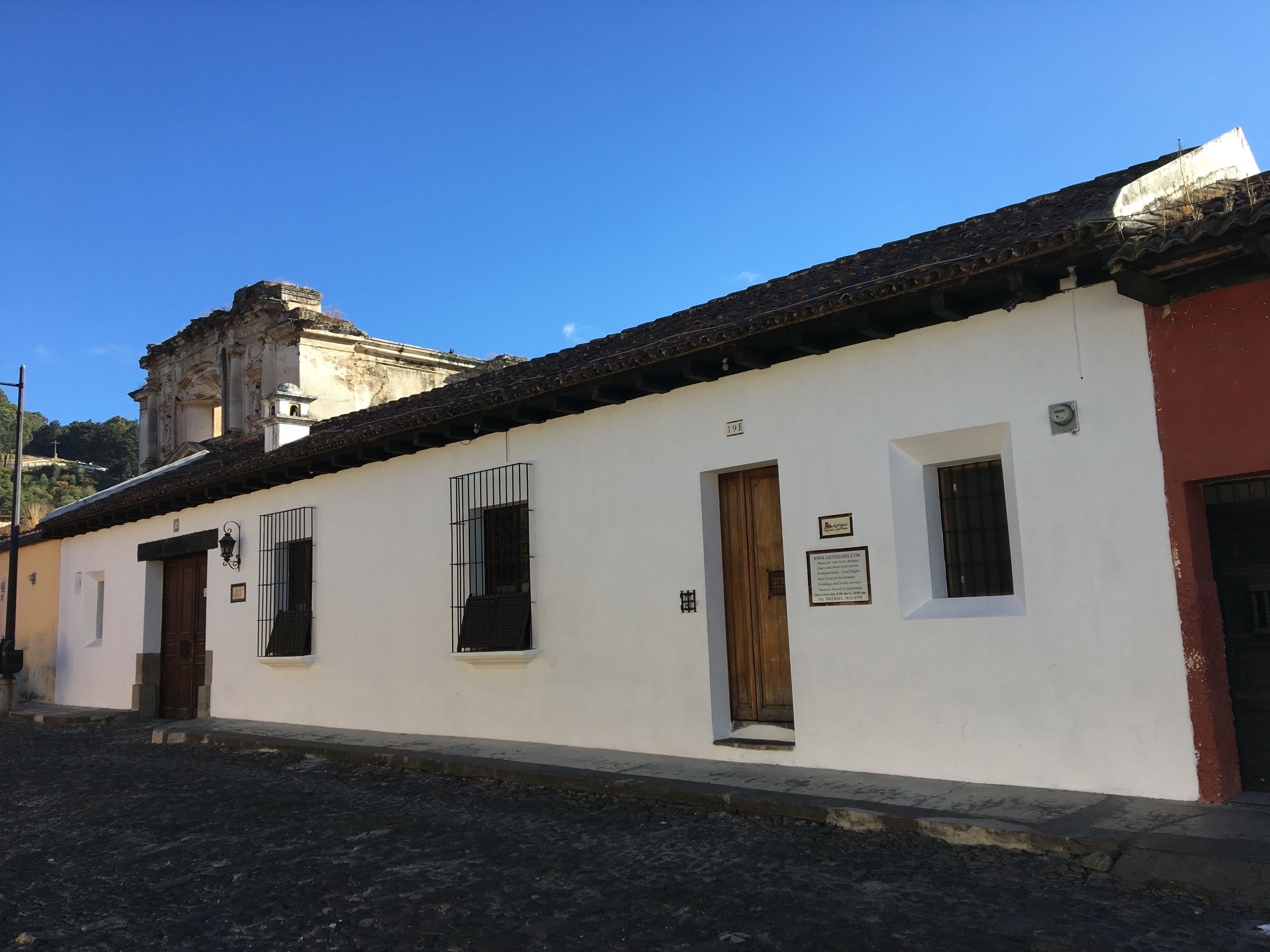 A white building with a tiled roof, numbered 19E, featuring a wooden door and a small window with a metal grill.