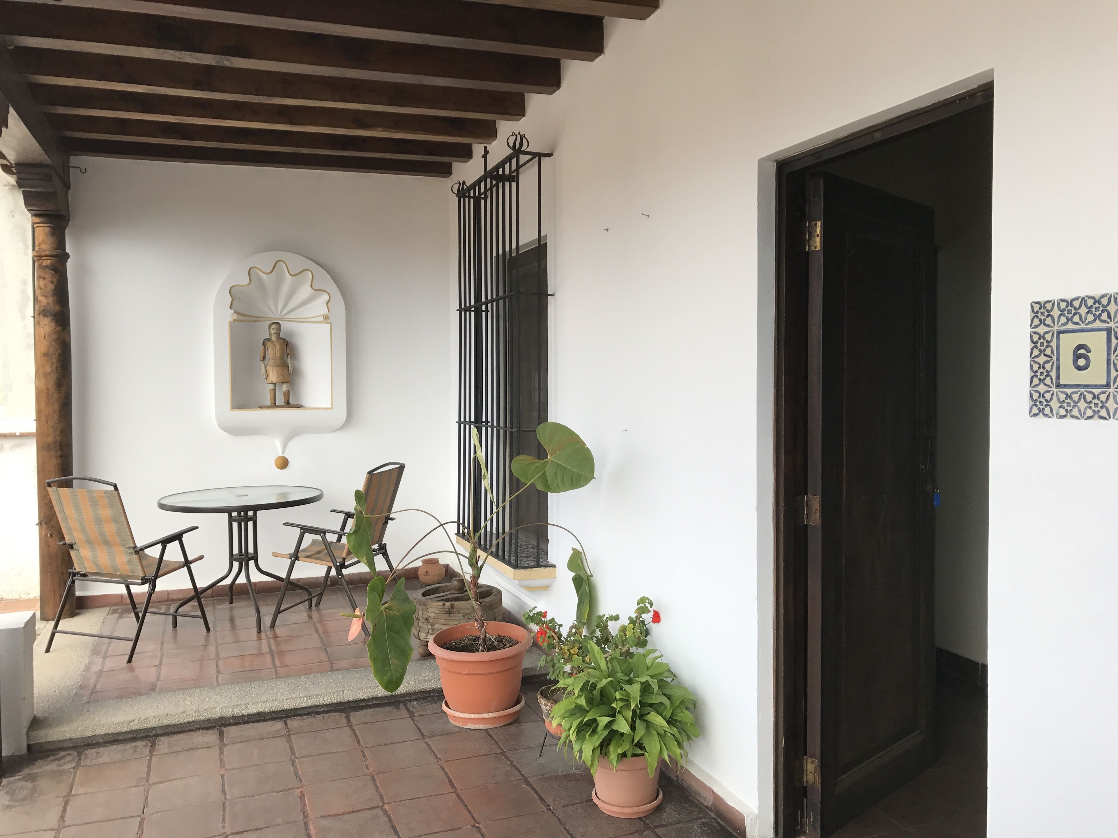 A patio area with a round table and two chairs, potted plants, a decorative mirror, and a door with a number plaque.