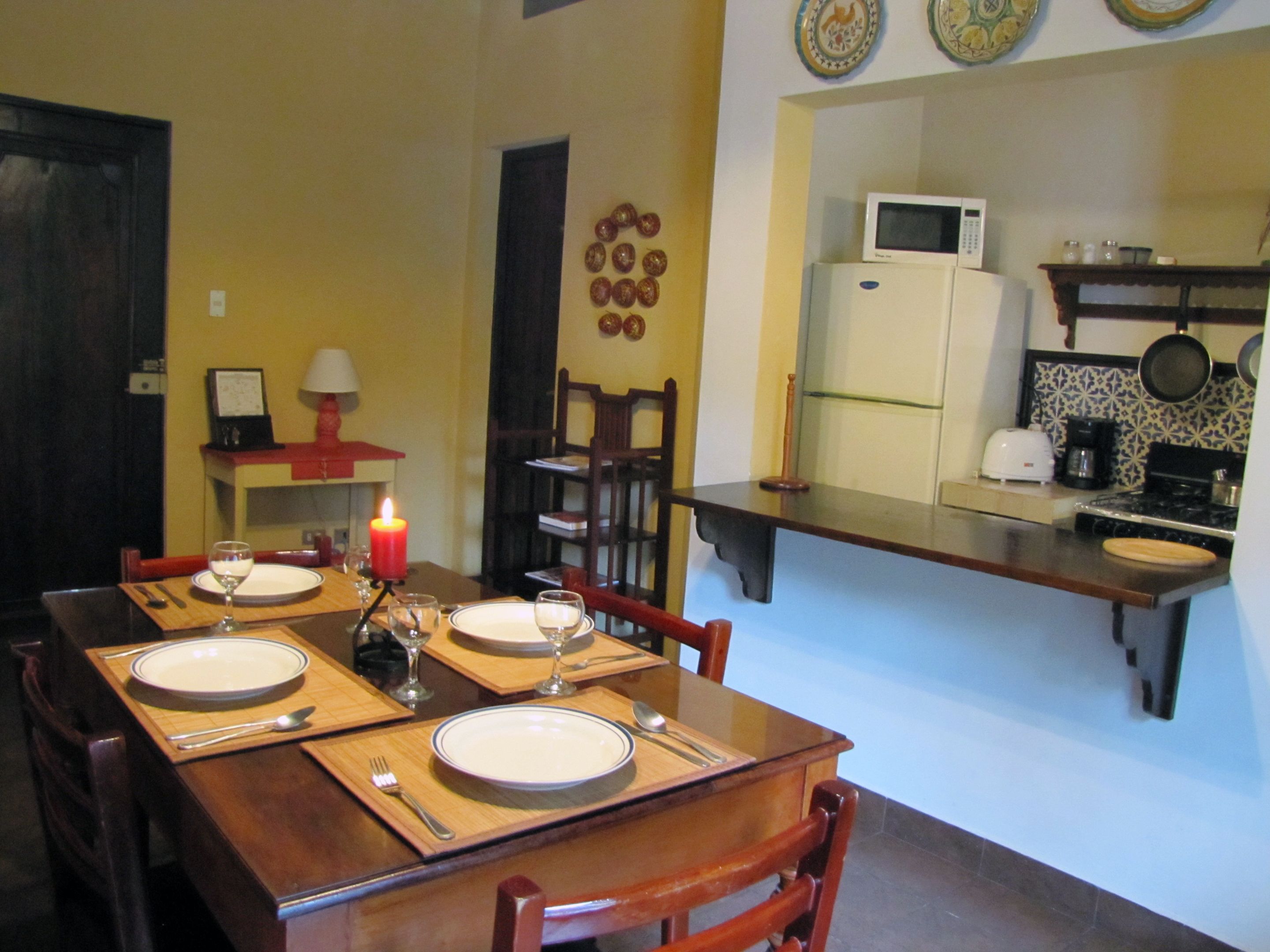 A dining area with a wooden table set for four, a lit candle, and a kitchen area with a microwave and stove.