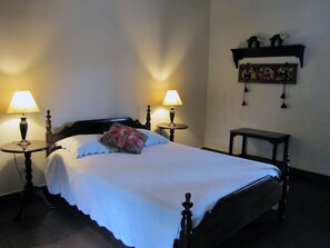 A neatly made bed with a floral pillow, two bedside lamps, a small table, and a shelf with decorative items.
