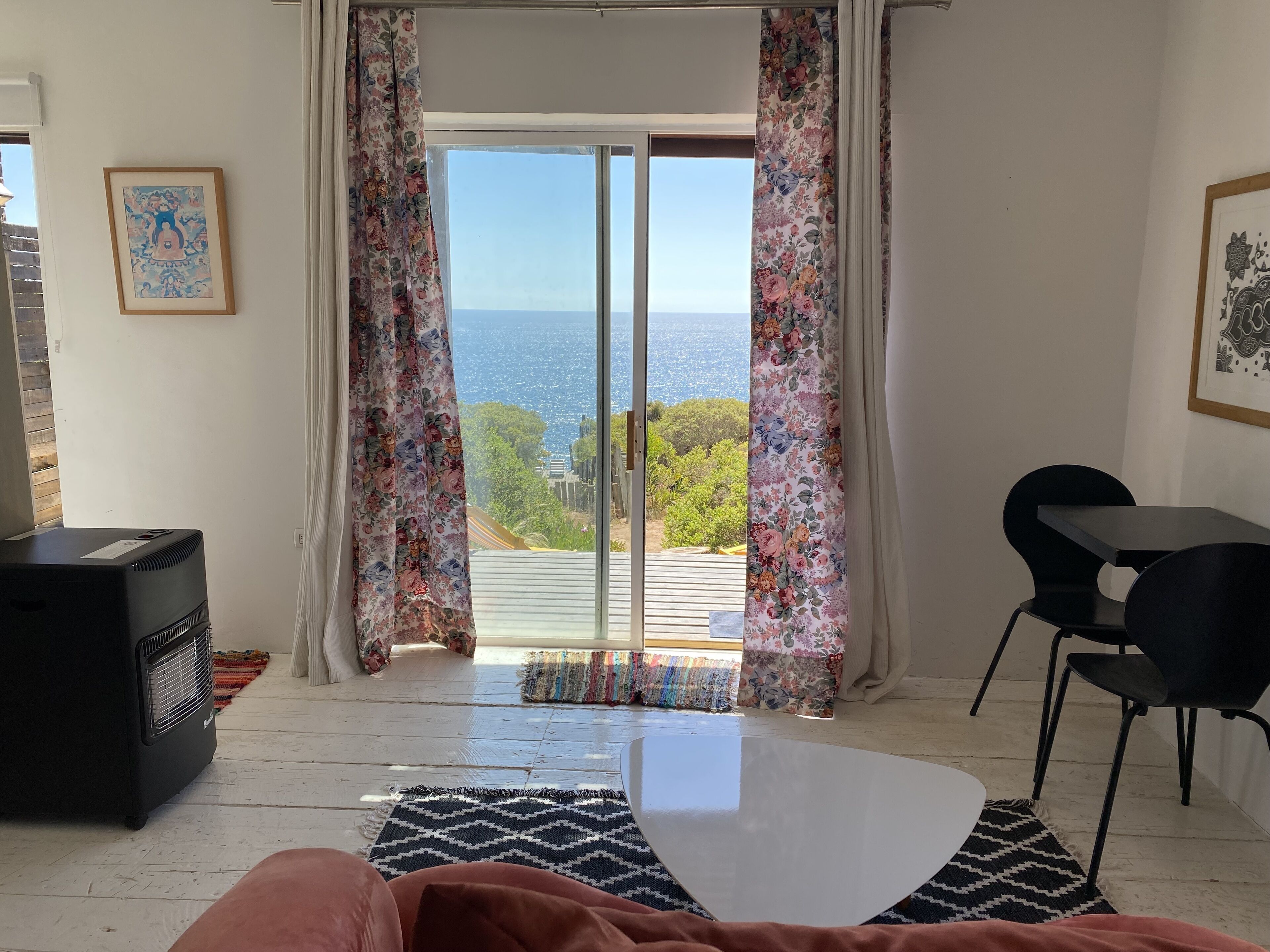 Una habitación con un amplio ventanal corredero que da a un balcón con vistas al mar. Hay un calefactor negro, una mesa blanca y una silla negra.