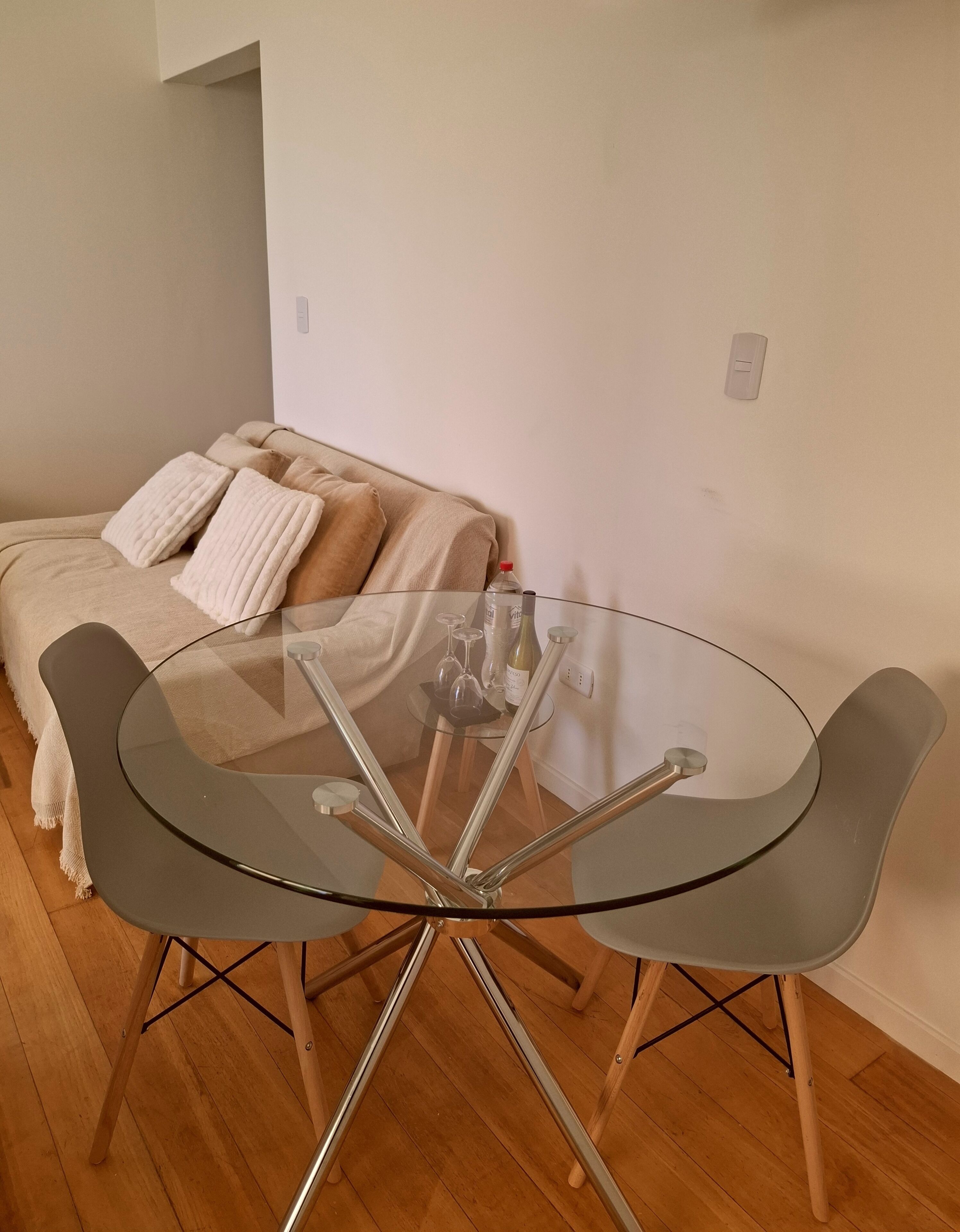 Ein runder Glastisch mit Metallbeinen, ein beiges Sofa mit Kissen und eine weiße Wand.