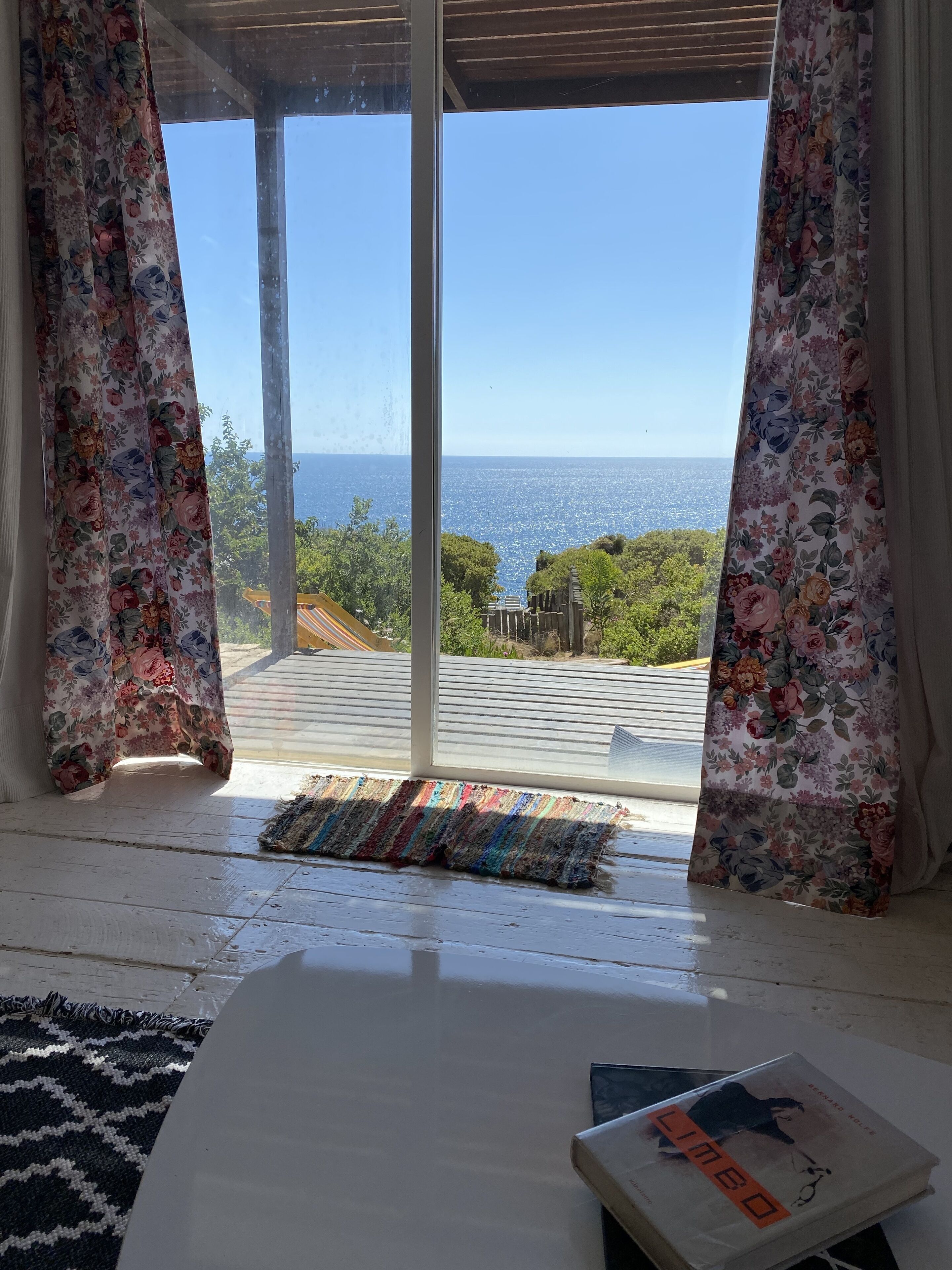 Une pièce dotée d’une grande fenêtre, de rideaux à motifs floraux, d’un plancher en bois, d’une table blanche et d’un livre intitulé « Limeo ».