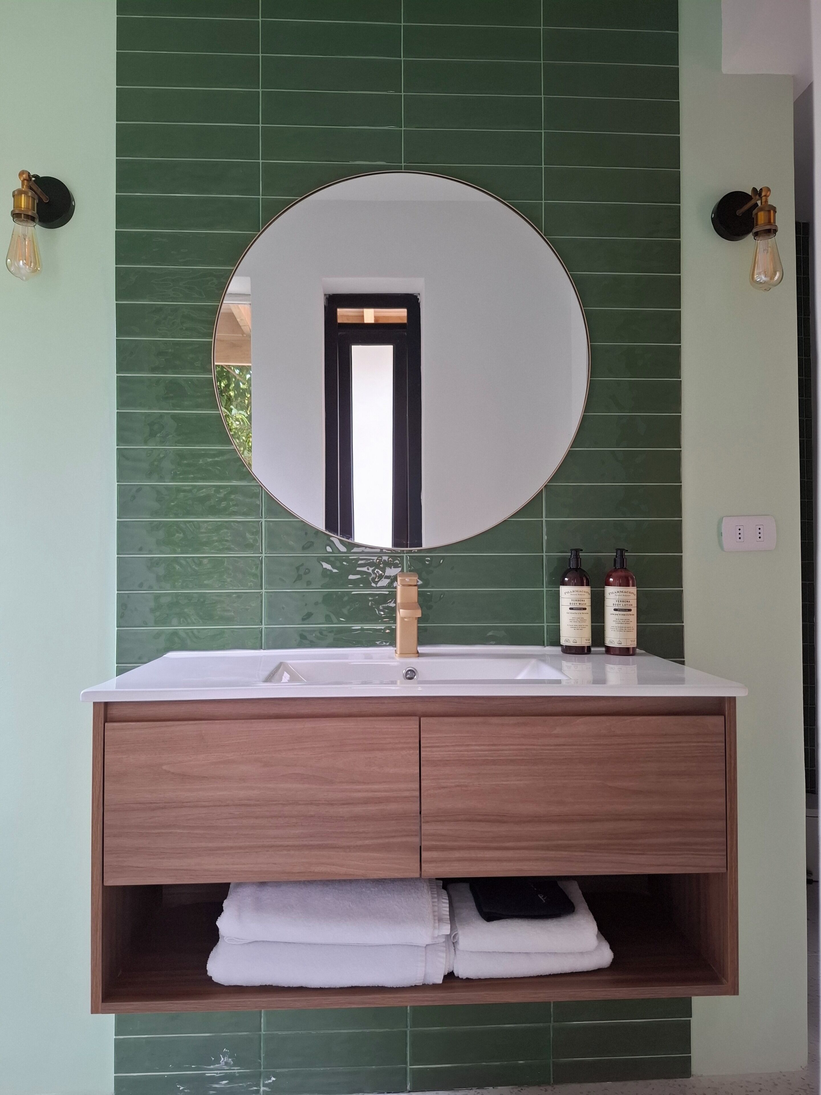 A bathroom with a round mirror, a white sink, and a green tiled wall.
