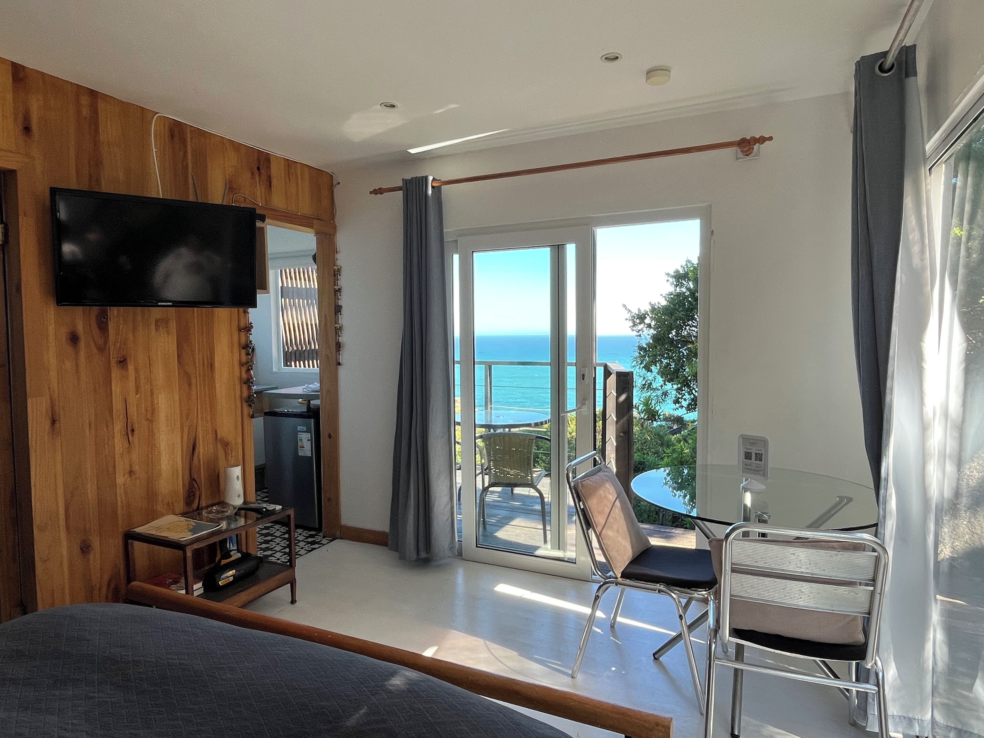 Un dormitorio con televisión, cama, escritorio y silla. Hay un balcón con vista al océano.