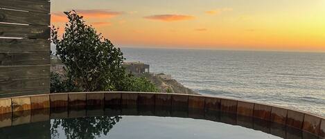 A pool with a wooden deck and a view of the ocean at sunset.