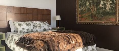 Un dormitorio con cabecera de madera, cama con ropa de cama floral, un plaid de piel, mesita de noche, lámpara y un cuadro enmarcado en la pared.