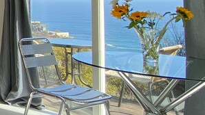 A balcony with a glass table and chairs, a vase with flowers, and a view of the ocean.