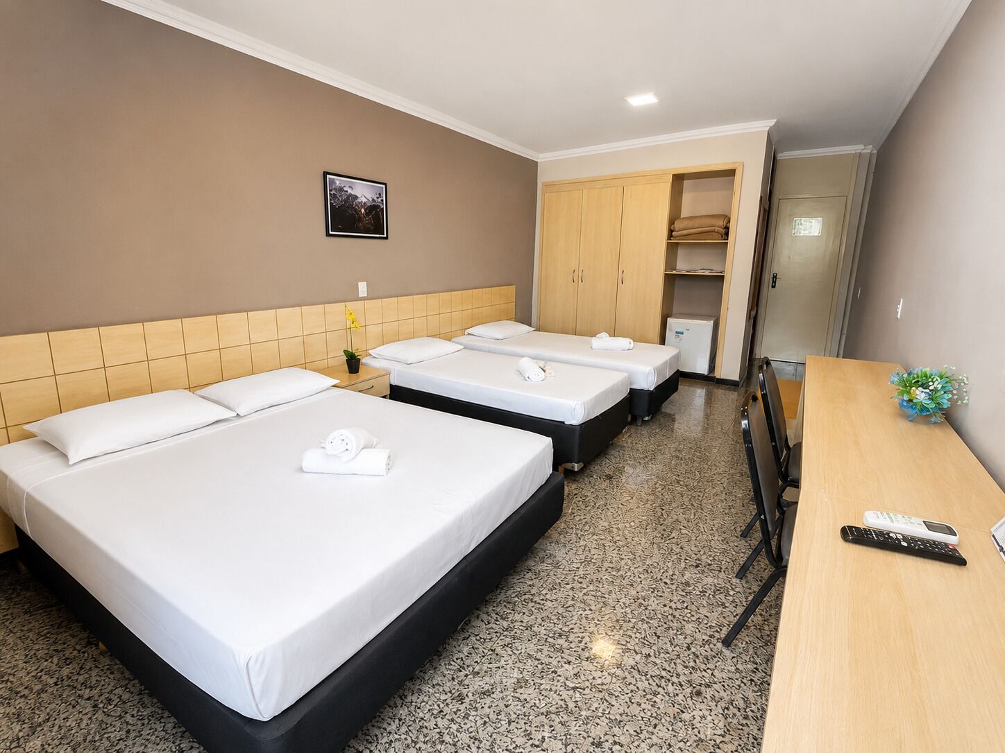 Habitación de hotel con tres camas, un escritorio, un refrigerador pequeño y una puerta que da a otra habitación.