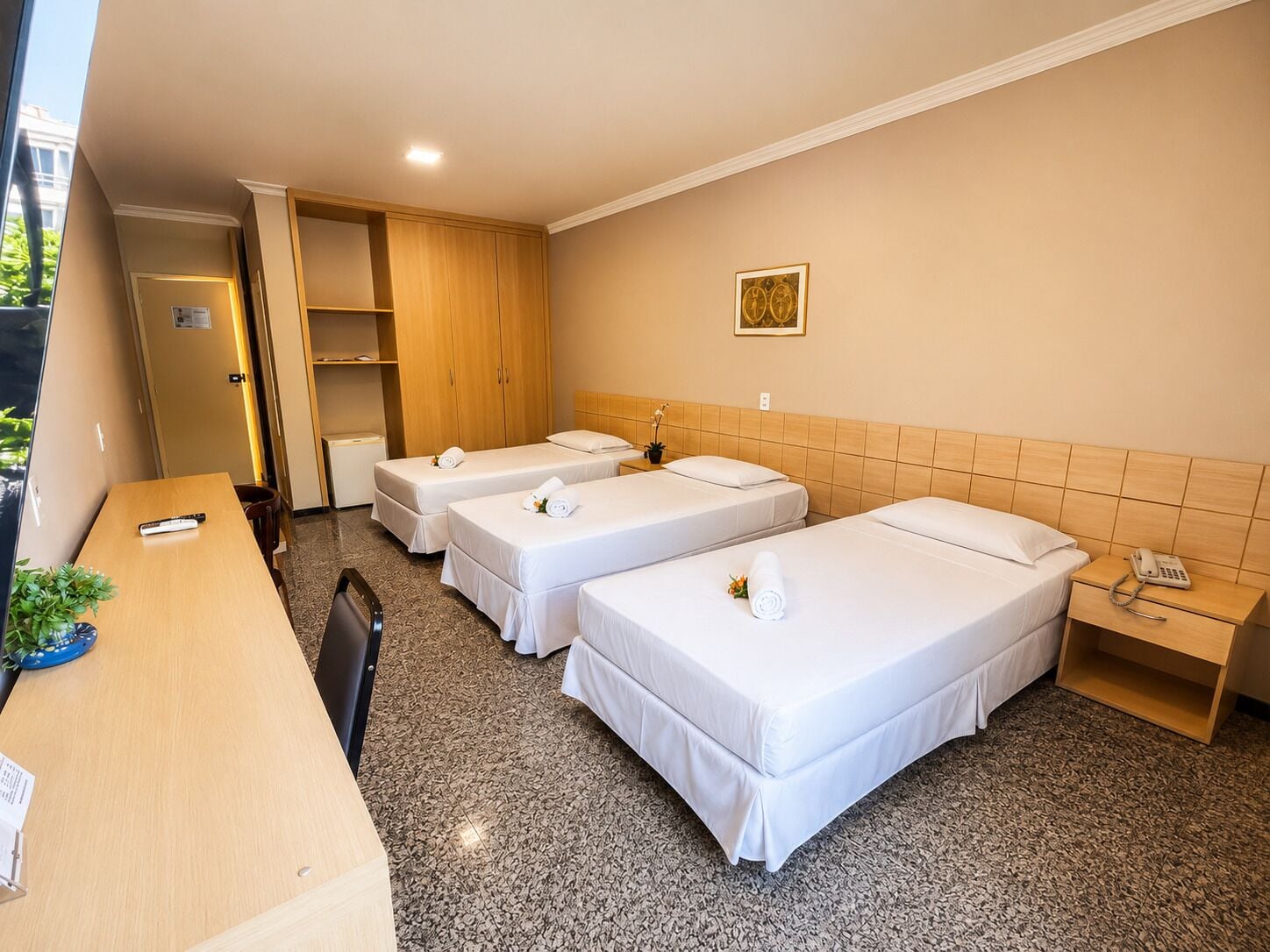 Habitación de hotel con tres camas individuales, un escritorio con silla, teléfono y una planta pequeña.