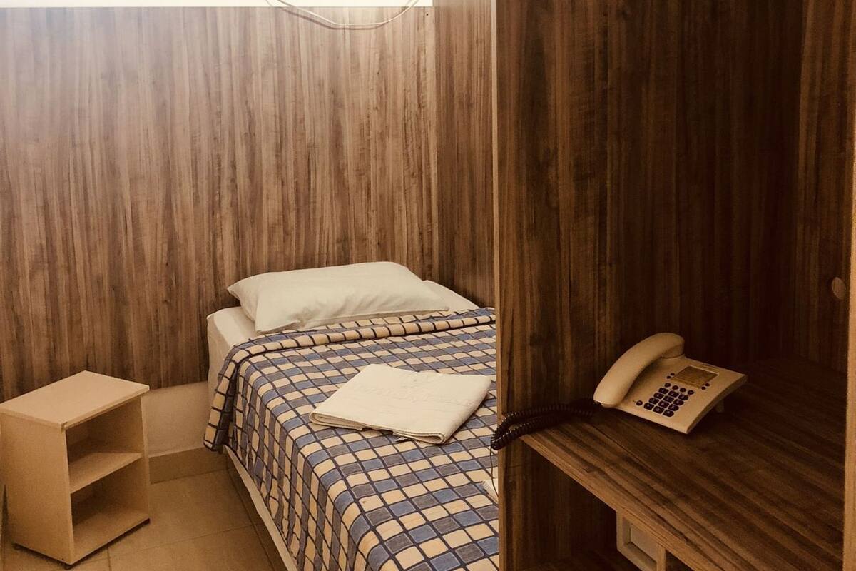 A small, compact room with a bed, a bedside table, a hanging clothes rack, and a wall-mounted telephone.