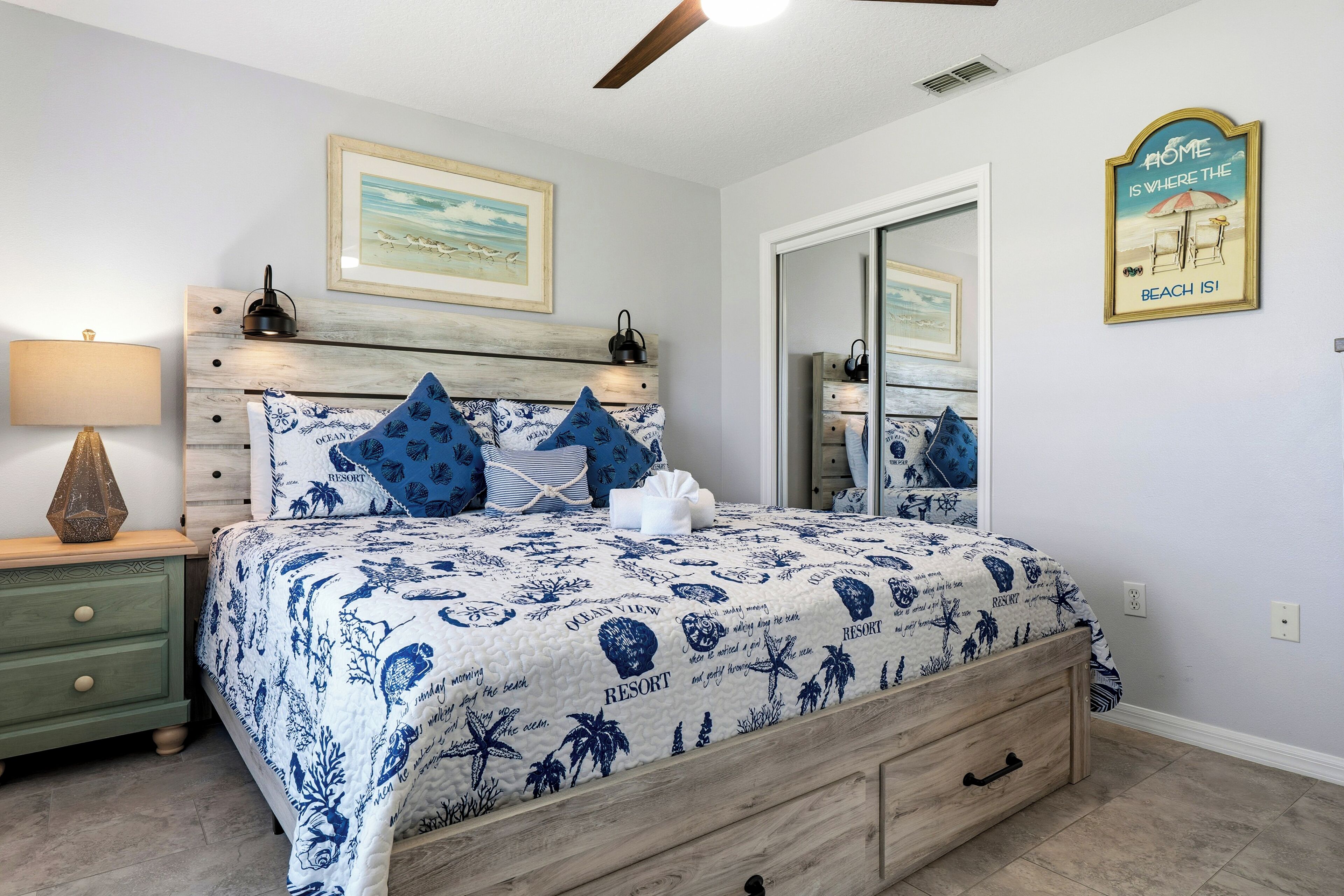 A bedroom with a bed, wooden headboard, decorative pillows, a nightstand, a lamp, a framed picture, and a wall sign.