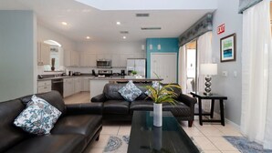 Una sala de estar moderna con un sofá, una mesa de centro y una planta. Se observa una cocina al fondo con armarios blancos y una pared azul decorativa.