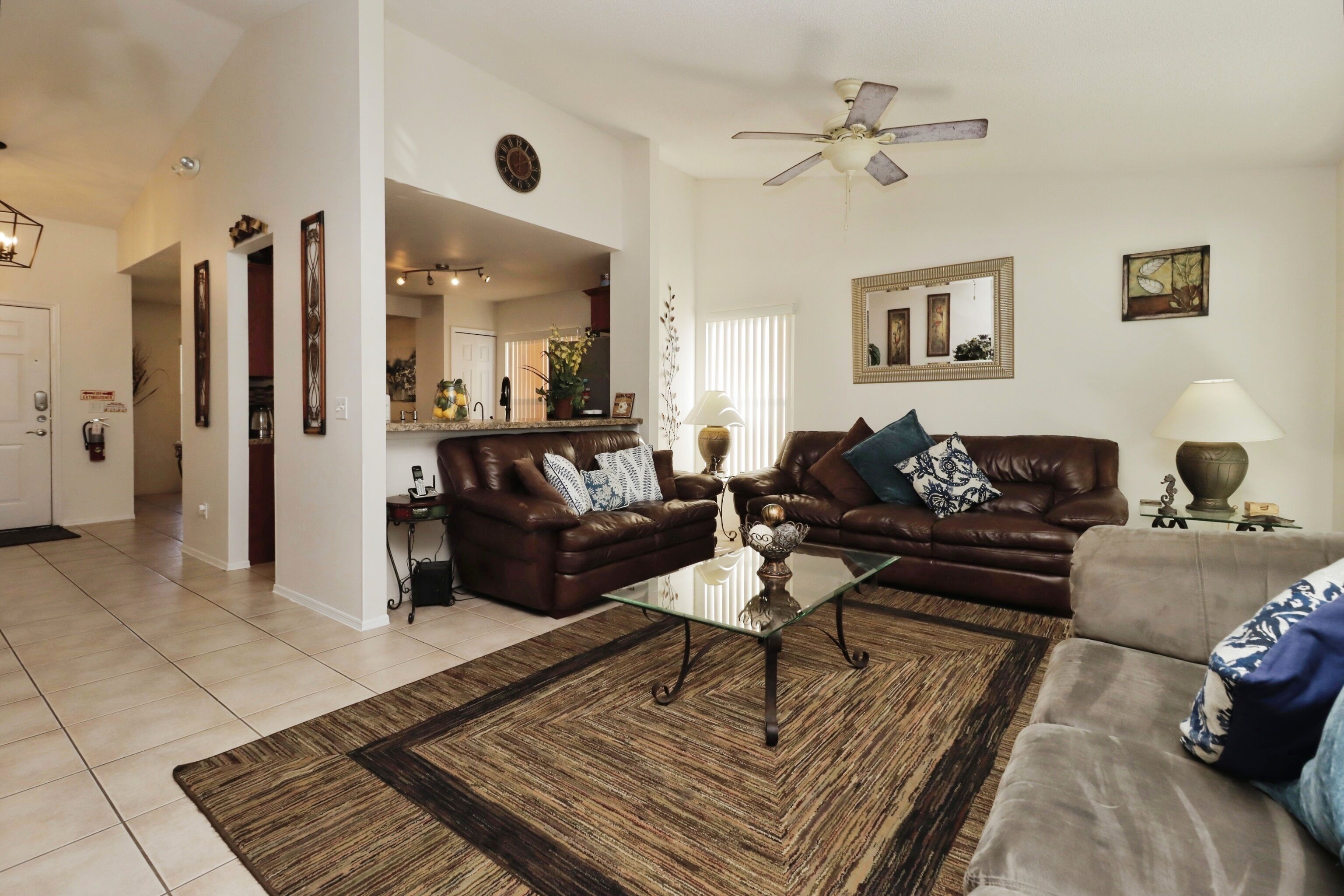 A spacious living room with a brown leather sofa, a glass coffee table, and a grey sofa. There is a ceiling fan, a large mirror, and a decorative vase on the coffee table.