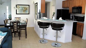 Una cucina moderna con mobili in legno, un frigorifero nero e un piano di lavoro in granito. Sono presenti due sgabelli da bar neri e un tavolo da pranzo con sedie.