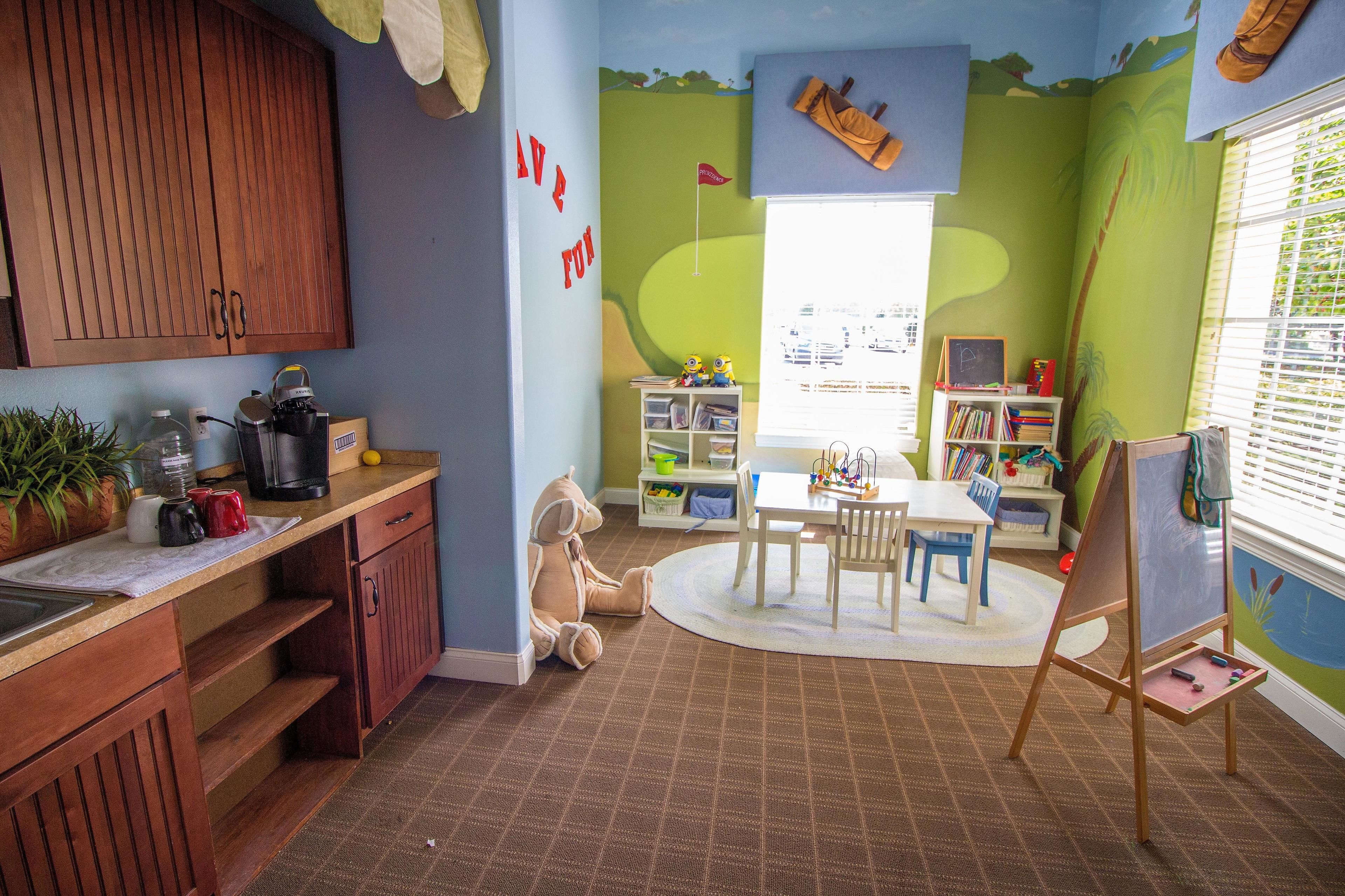 A child's playroom with a wooden cabinet, a desk with a coffee maker, a chair, a small table, and a bookshelf.