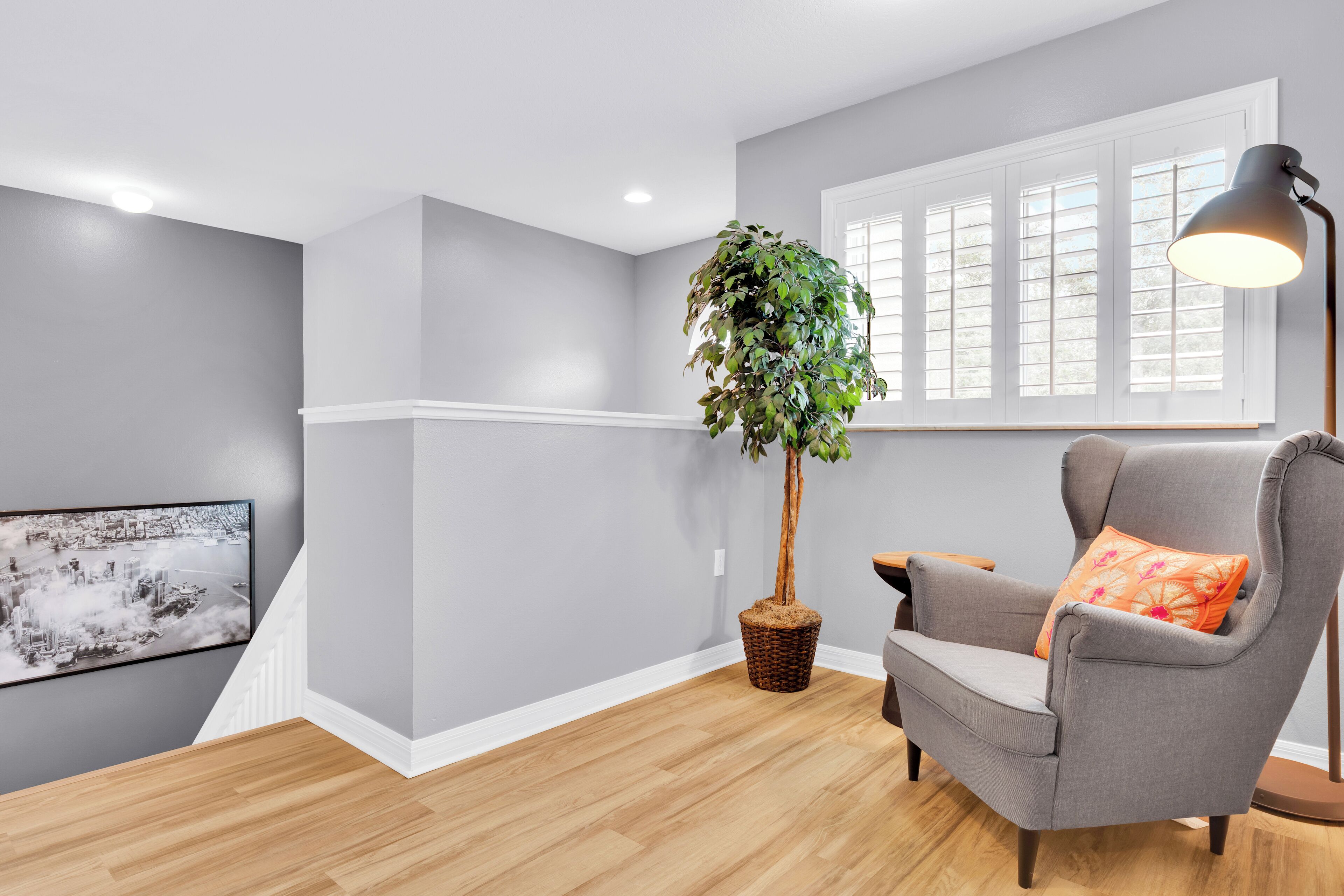A room with grey walls, a wooden floor, a grey armchair with an orange pillow, a floor lamp, a large window with blinds, and a wall-mounted TV.