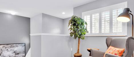 A room with grey walls, a wooden floor, a grey armchair with an orange pillow, a floor lamp, a large window with blinds, and a wall-mounted TV.