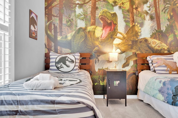 Une chambre à coucher avec une fresque murale sur le thème des dinosaures, une literie à rayures et une table de chevet avec une lampe.