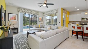 A modern living room with a sectional sofa, a coffee table, and a dining area with chairs. There is a ceiling fan, a kitchen with cabinets, and a large window offering a view of the outdoors.