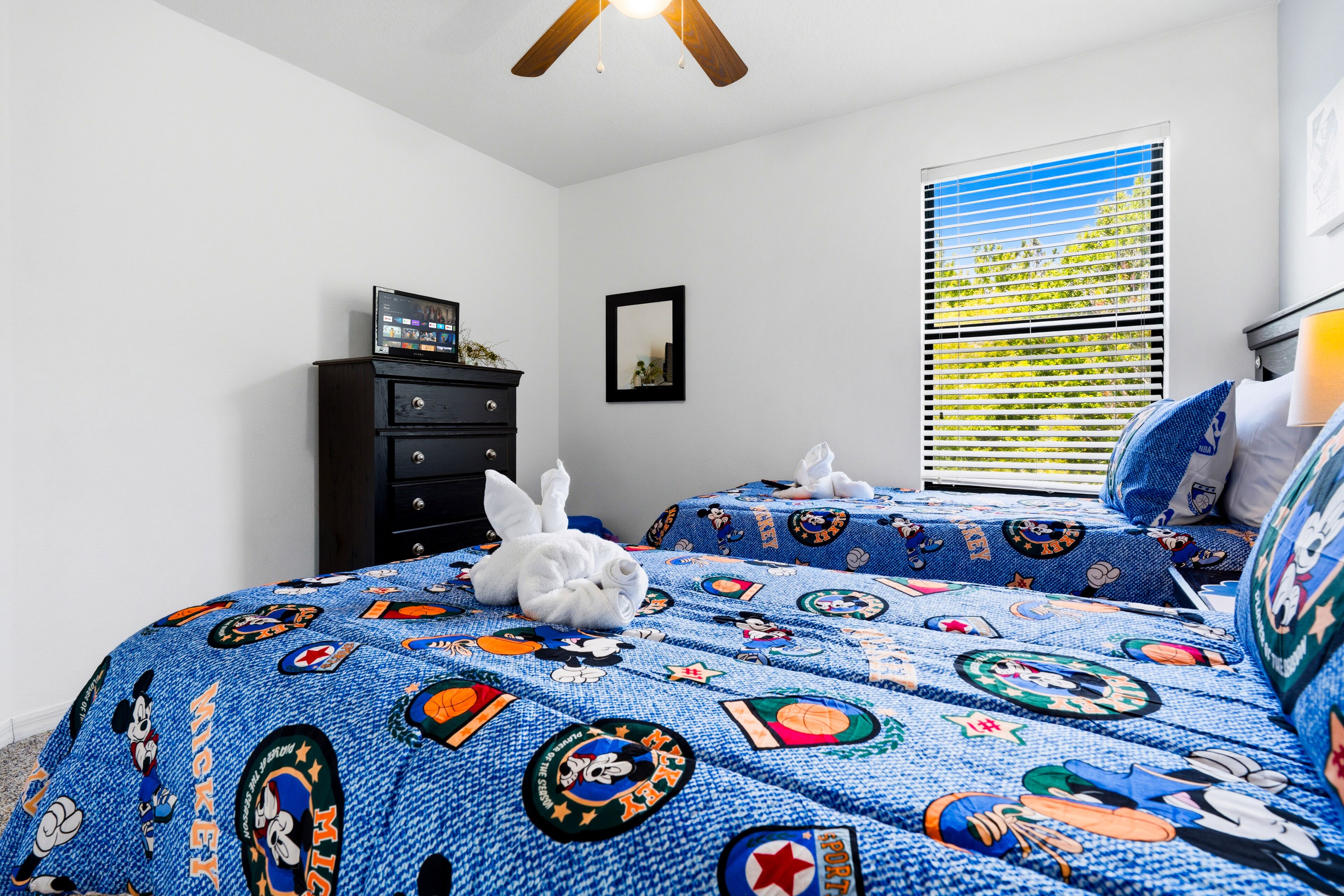A bedroom with a bed featuring a sports-themed quilt, a dresser, a window with blinds, and a ceiling fan.