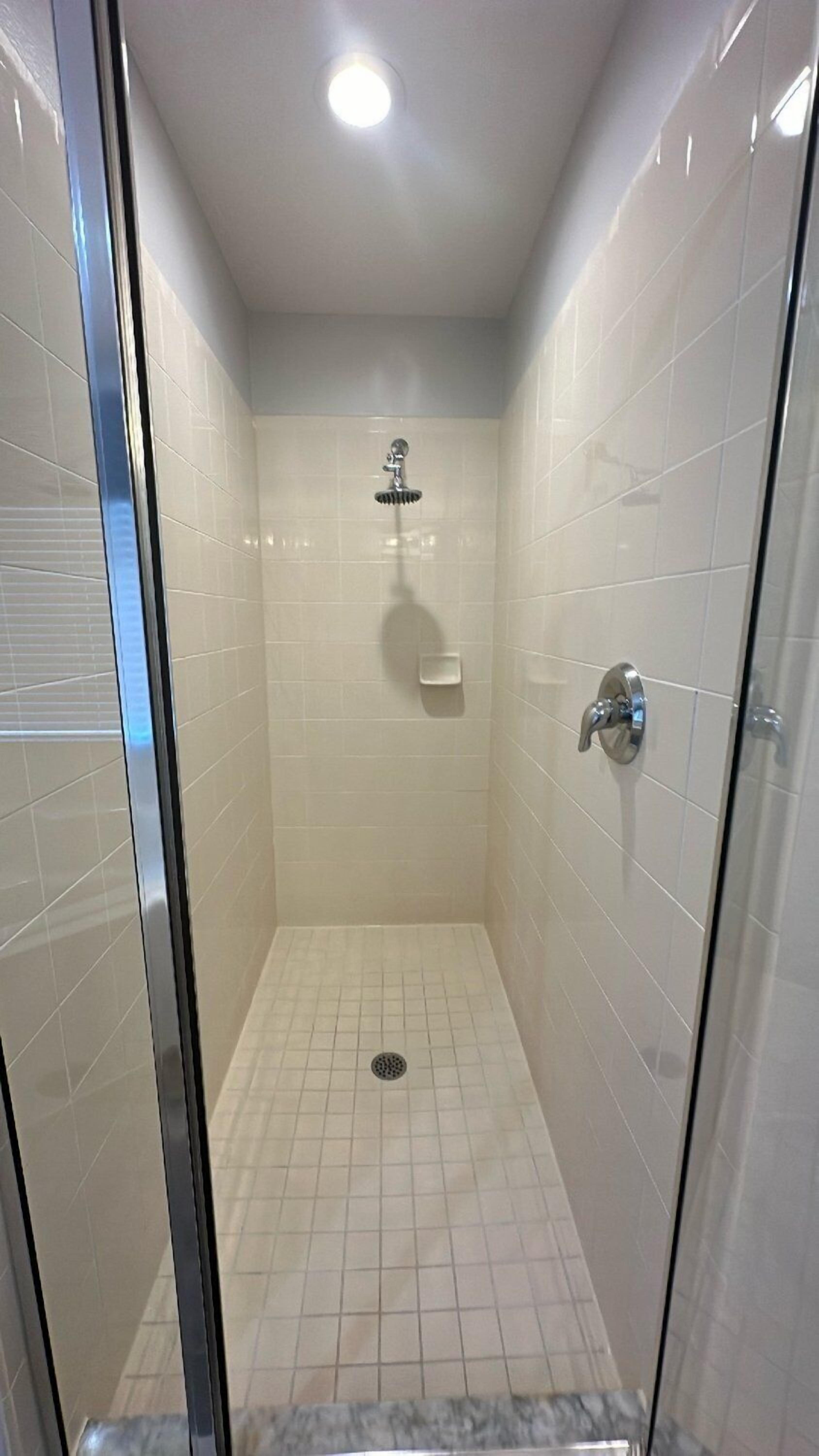 A glass-enclosed shower stall with white tiled walls and floor, a showerhead, a soap dish, and a drain.