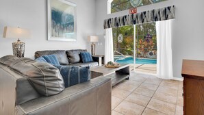 A modern living room with a leather sofa, a coffee table, and a pool view through large glass doors. - Allamanda Holiday Home 4 Bedroom (Kissimmee)