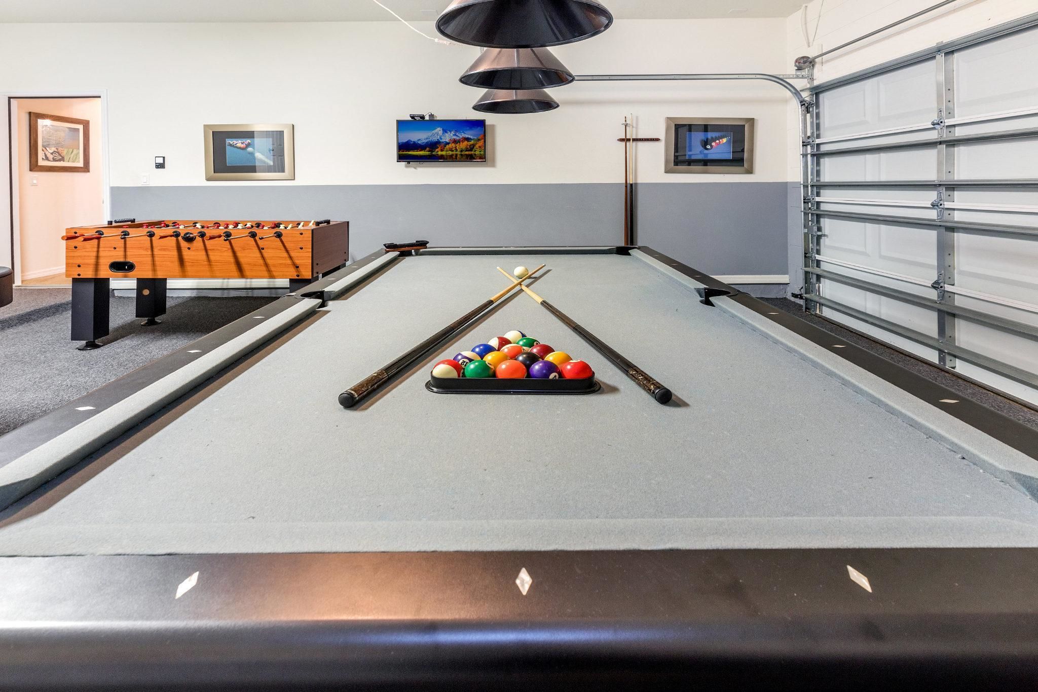A pool table with a cue stick and balls, a foosball table, and a garage door.