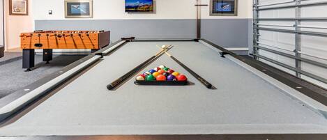 A pool table with a cue stick and balls, a foosball table, and a garage door.