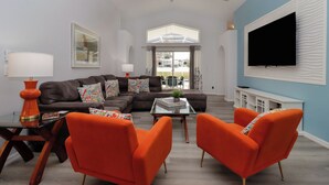 Een moderne woonkamer met een hoekbank, oranje fauteuils, een glazen salontafel en een flatscreen televisie aan de muur.