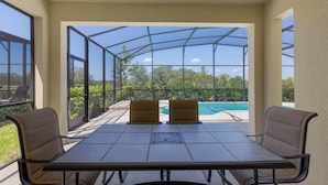 Une terrasse couverte, dotée d’une table et de chaises, qui offre une vue sur une piscine et un espace verdoyant.