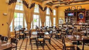 Un restaurant doté de nombreuses tables et chaises, de grandes fenêtres avec des rideaux, d’un lustre et d’un coin bar avec un téléviseur.