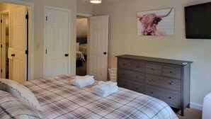Un dormitorio con colcha escocesa, cómoda, televisor montado y puerta que da a otra habitación.