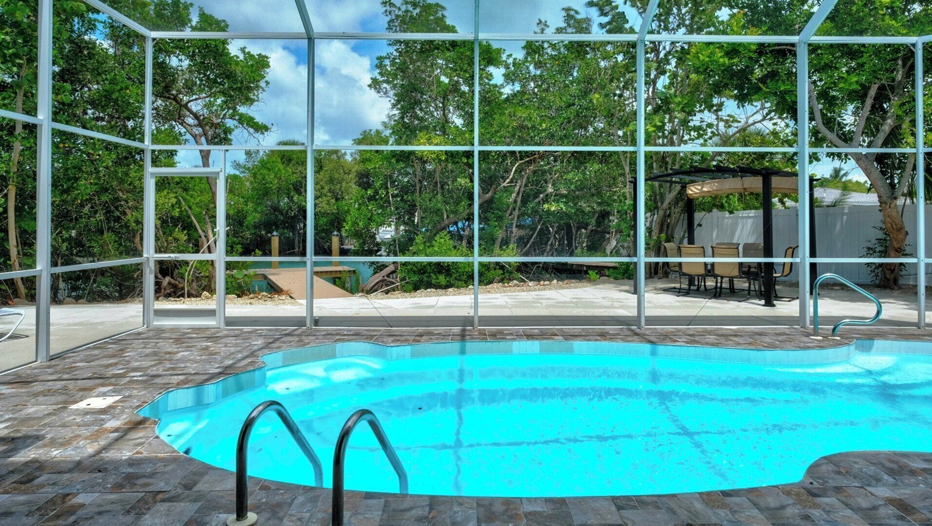 An indoor swimming pool with clear glass walls, surrounded by a patio area with outdoor furniture.