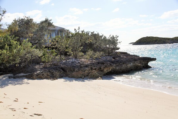 Une plage avec une maison à moitié dissimulée par la végétation, un rivage rocheux et une mer calme.