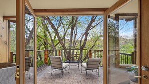 Une porte vitrée ouverte donne sur un balcon où se trouvent deux chaises et une table, le tout entouré d’une terrasse en bois et offrant une vue sur les arbres.