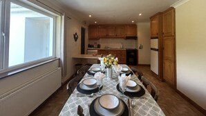 A dining area with a table set for four, kitchen cabinets, a refrigerator, and a window. - Waterside Cottage stunning sea & mountain views. (Connemara)
