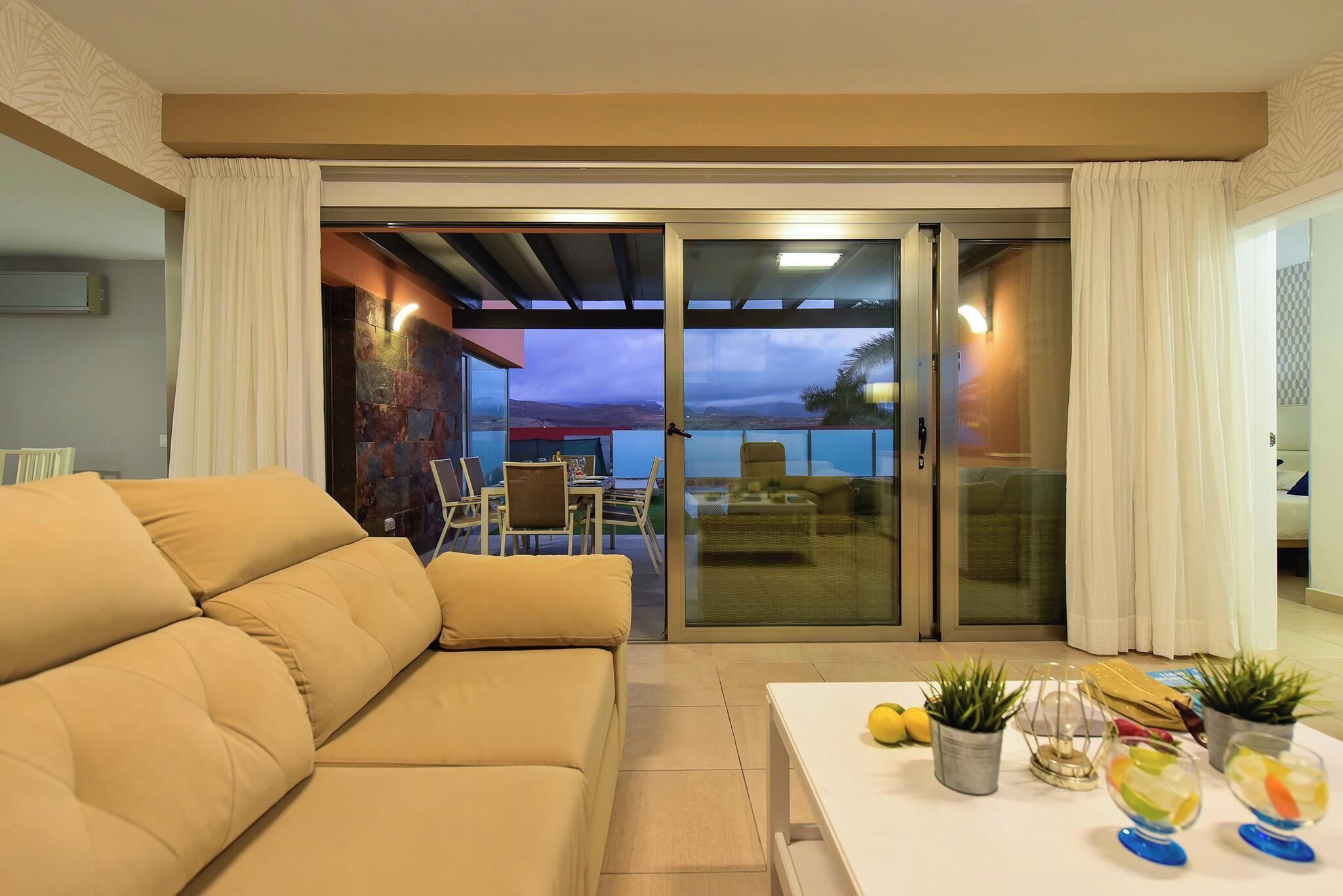 A modern living room with a beige sofa, a white coffee table with decorative items, and a sliding glass door leading to an outdoor area with a dining table and chairs.