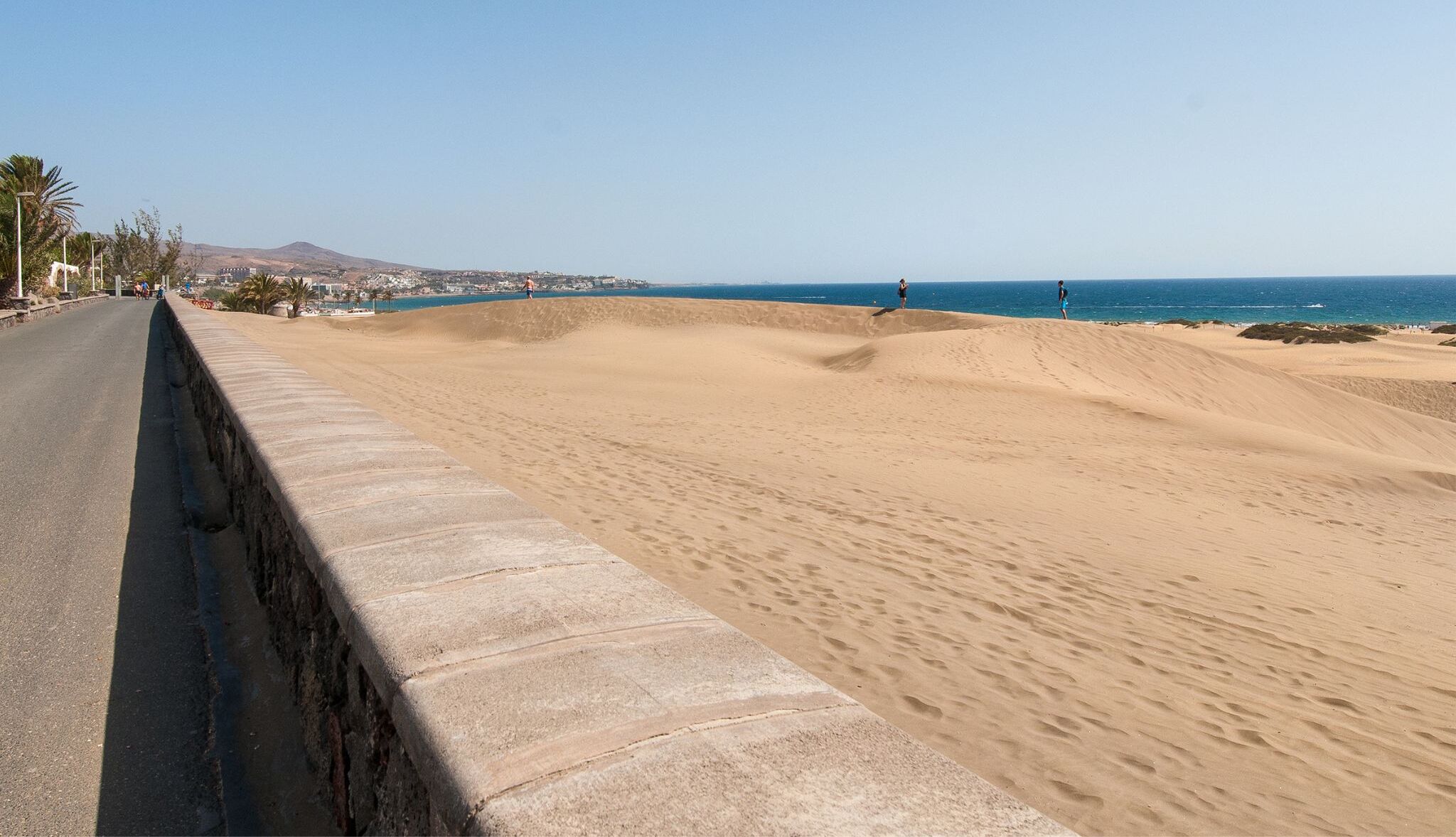 A coastal road with a sandy dune landscape, a clear sky, and distant buildings.
