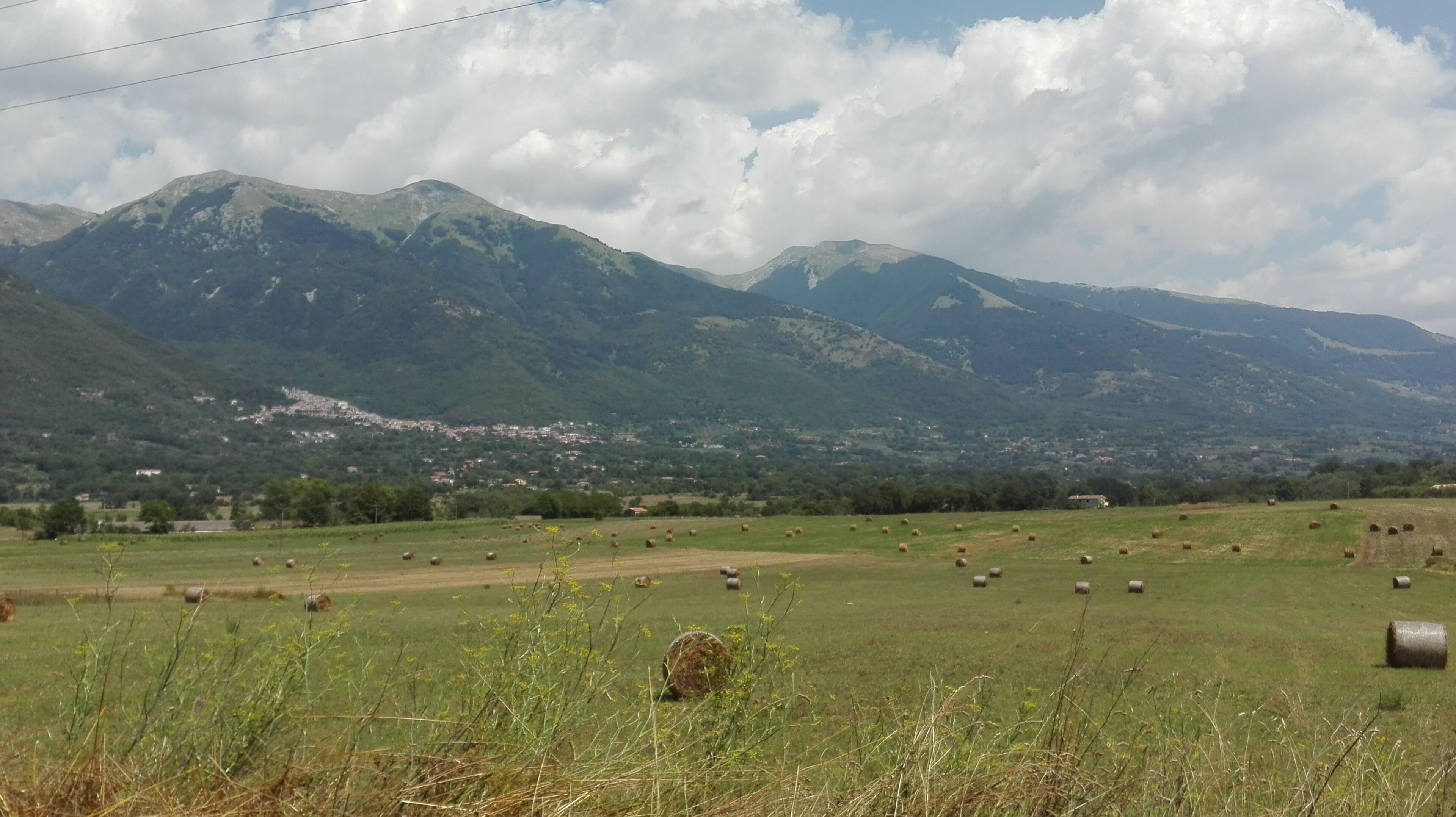 Paesaggio rurale con dolci colline, un campo di balle di fieno e un paese in lontananza.