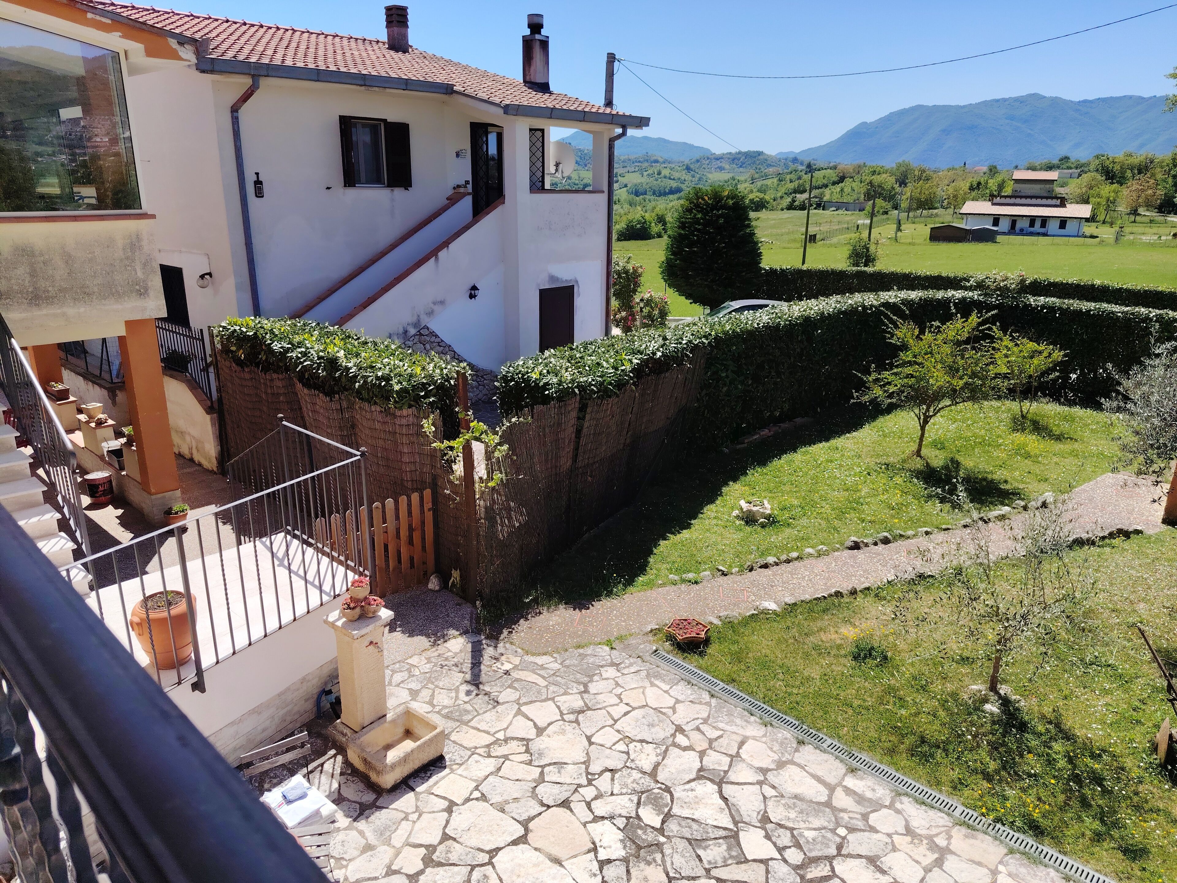 Una casa con patio in pietra, recinzione in legno e vista su un paesaggio verde con montagne.